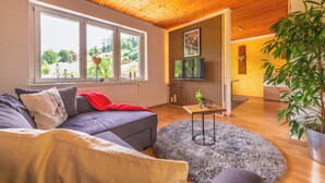 Ein modernes Wohnzimmer mit Holzbalkendecke, einem grauen Sofa, einem runden Teppich und einem großen Fenster mit Blick auf Grünflächen.