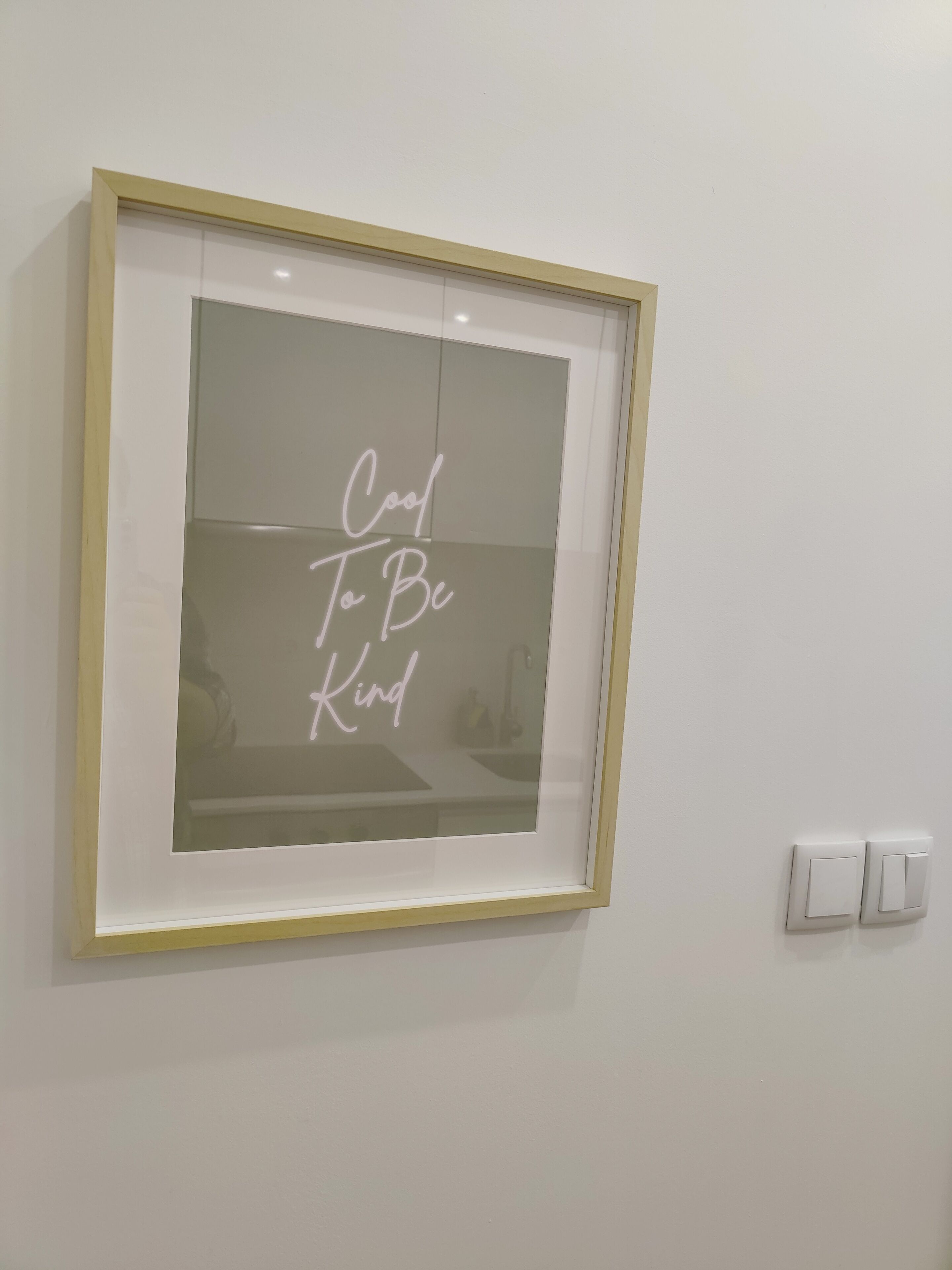 A framed sign with the text "Cool To Be Kind" on a white wall.