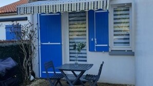 A small outdoor seating area with a table and chairs, a blue door, and a window with shutters. - Comfortable Cottage 4 Pers, Beach 200m, Private Parking (La Faute-sur-Mer)