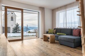 Ett modernt vardagsrum med trägolv, en grå soffa med kuddar, ett träbord och en stor skjutdörr i glas som leder ut till ett snötäckt utomhusområde med en kyrka och berg i bakgrunden.