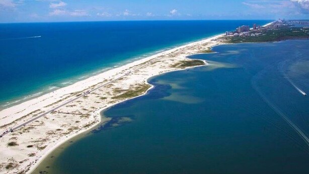 An aerial view of a coastal area with a long sandy beach, a clear blue ocean, and a small settlement with buildings near the shore. - Exclusive 2850 sf Pool Home near Perdido Beaches & NAS, Heated Pool, 2 Masters! (Pensacola)