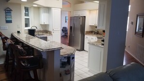 A kitchen with a central island, white cabinets, a stainless steel refrigerator, and a dining area with bar stools. - Exclusive 2850 sf Pool Home near Perdido Beaches & NAS, Heated Pool, 2 Masters! (Pensacola)