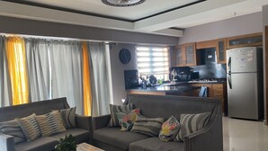 Een moderne woonkamer met een grijze zithoek, een houten bijzettafeltje met een plant, en een keuken met een koelkast, magnetron en fornuis.