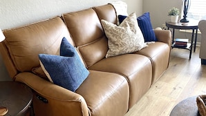 A tan leather sofa with blue and white cushions, a small round table with a lamp, and a view of a window with blinds.