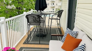 A balcony with a white railing, a black table with chairs, a white sofa with striped pillows, and a potted plant with pink flowers.