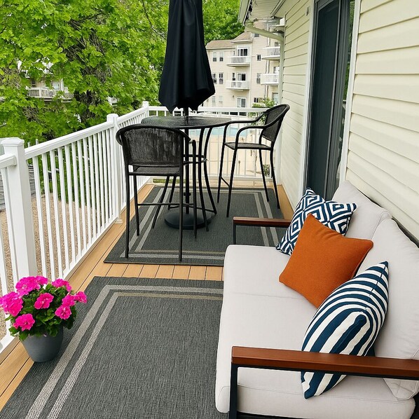 A balcony with a white railing, a black table with chairs, a white sofa with striped pillows, and a potted plant with pink flowers.