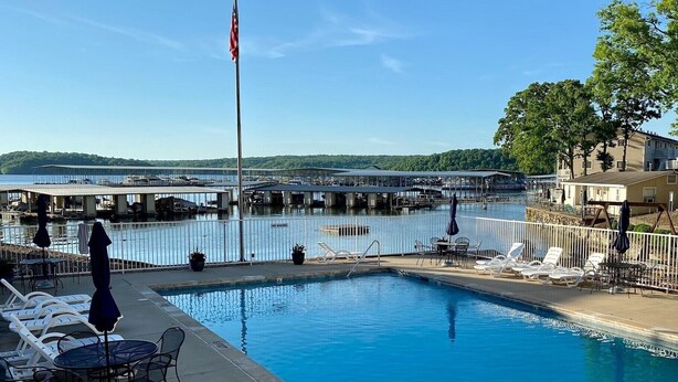 A swimming pool with lounge chairs and tables, a flagpole with an American flag, and a view of a marina with boats and a bridge in the background.