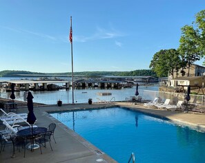 A swimming pool with lounge chairs and tables, a flagpole with an American flag, and a view of a marina with boats and a bridge in the background.