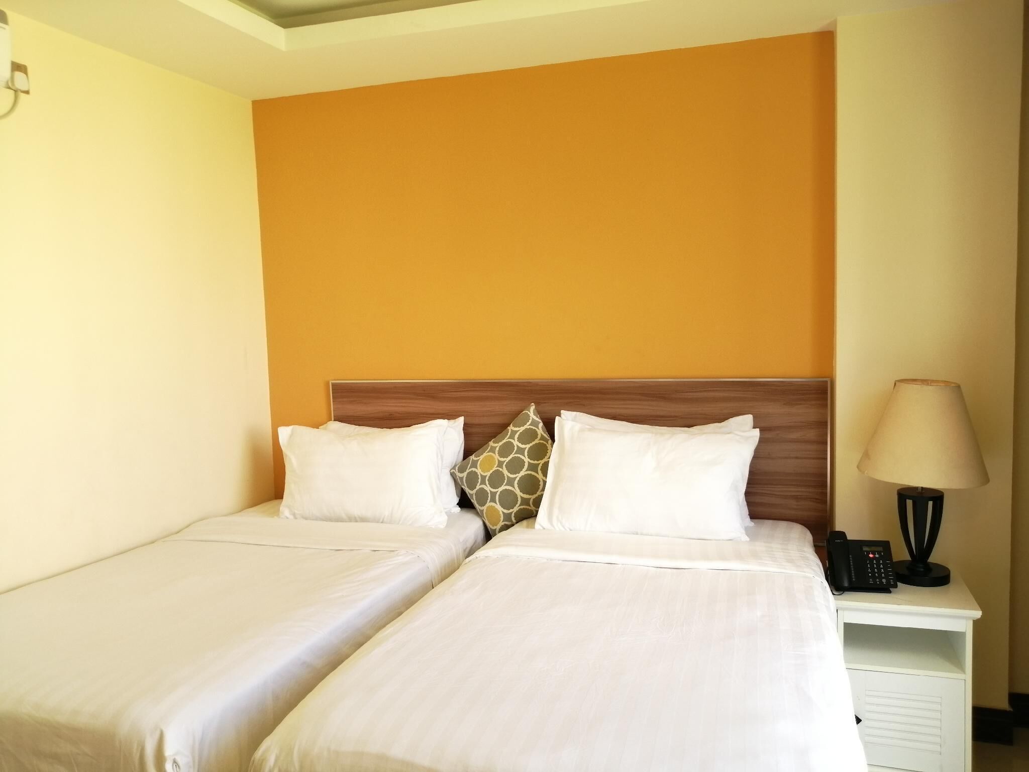 Habitación de hotel con dos camas, cabecero de madera, mesita de noche con lámpara y teléfono, y pared naranja lisa.