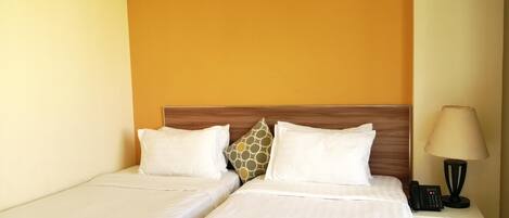 Habitación de hotel con dos camas, cabecero de madera, mesita de noche con lámpara y teléfono, y pared naranja lisa.