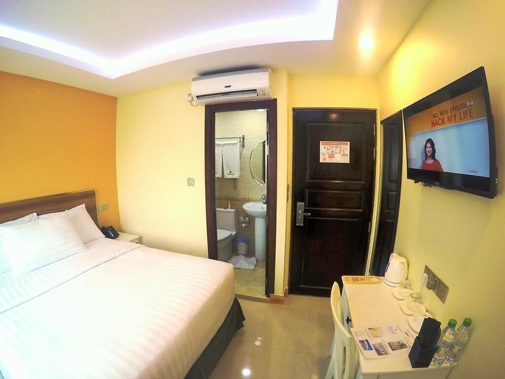 Quarto de hotel com cama, escrivaninha com telefone, espelho e televisão exibindo um programa.