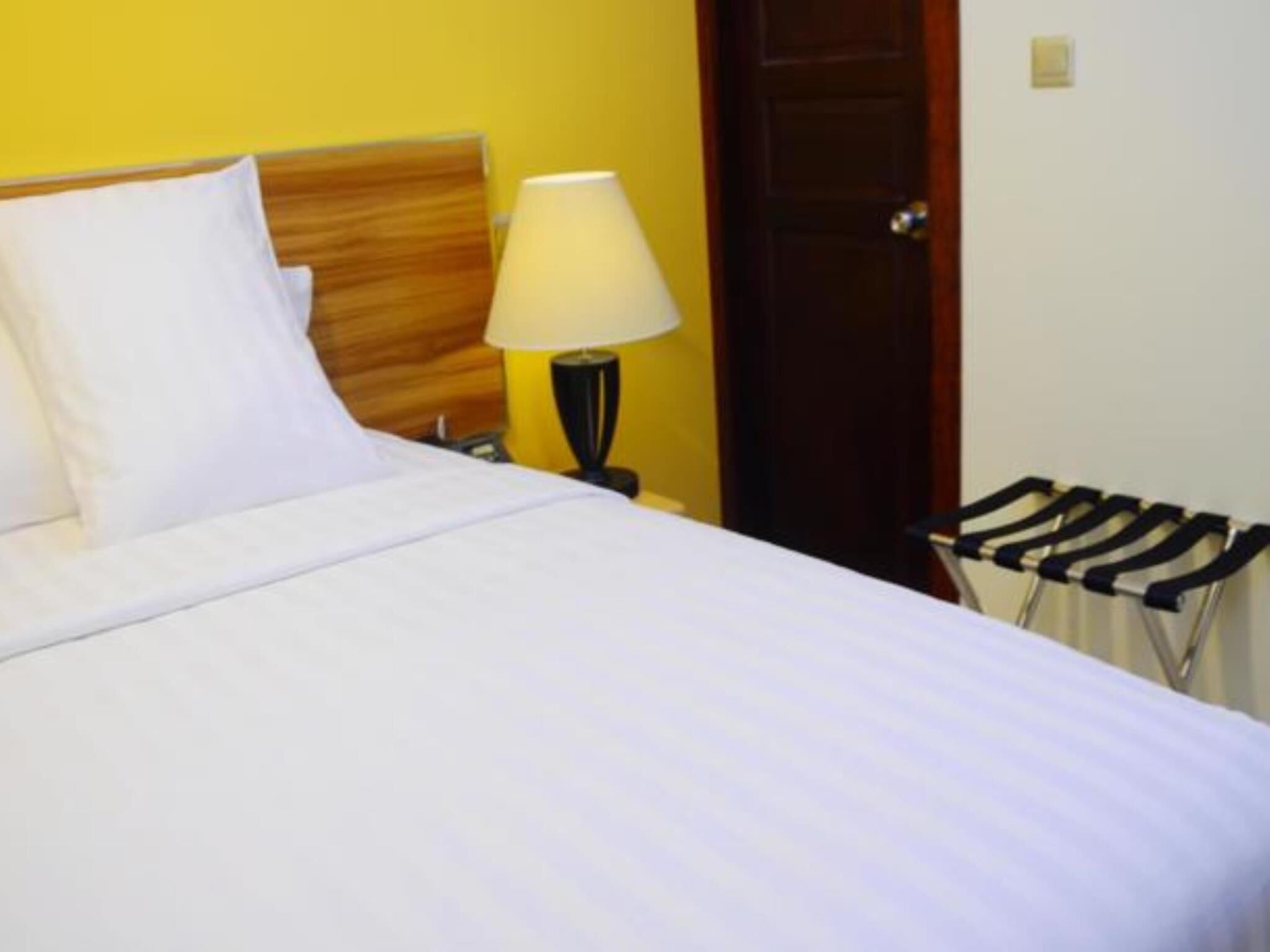 A neatly made bed with white linens, a wooden headboard, a bedside lamp, and a door in the background.