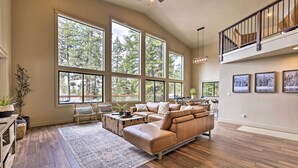 A spacious living room with large windows, a leather sofa, a wooden coffee table, and a staircase to an upper level. - McCall Family Retreat | Pool, Hot Tub & Bikes (McCall)