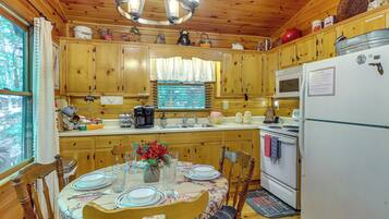 Une cuisine rustique aménagée dans une cabane en bois, avec une table à manger pour deux personnes, un réfrigérateur et une fenêtre avec des rideaux.