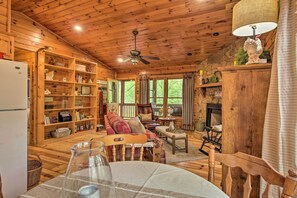 A cozy cabin interior with wooden walls, a fireplace, a dining area, and a large bookshelf.