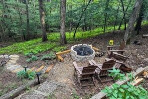 Un terrain de camping aménagé dans une forêt, avec un foyer, des chaises en bois et un panneau d’avertissement concernant les risques d’incendies de forêt.