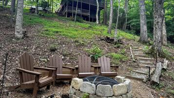 Un área boscosa con una terraza de madera, sillas Adirondack y un hoyo para el fuego de piedra.