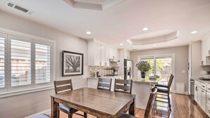 A spacious kitchen with white cabinets, a wooden dining table, and a view of the backyard through large windows. - Family Home in Walkable Area Near Silicon Valley! (San Jose)