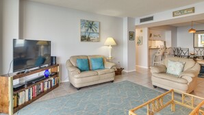 A living room with a flat-screen TV, a beige sofa, a coffee table, and a bookshelf. - Dolphin & Manatee Viewing From 15th Floor (Clearwater)