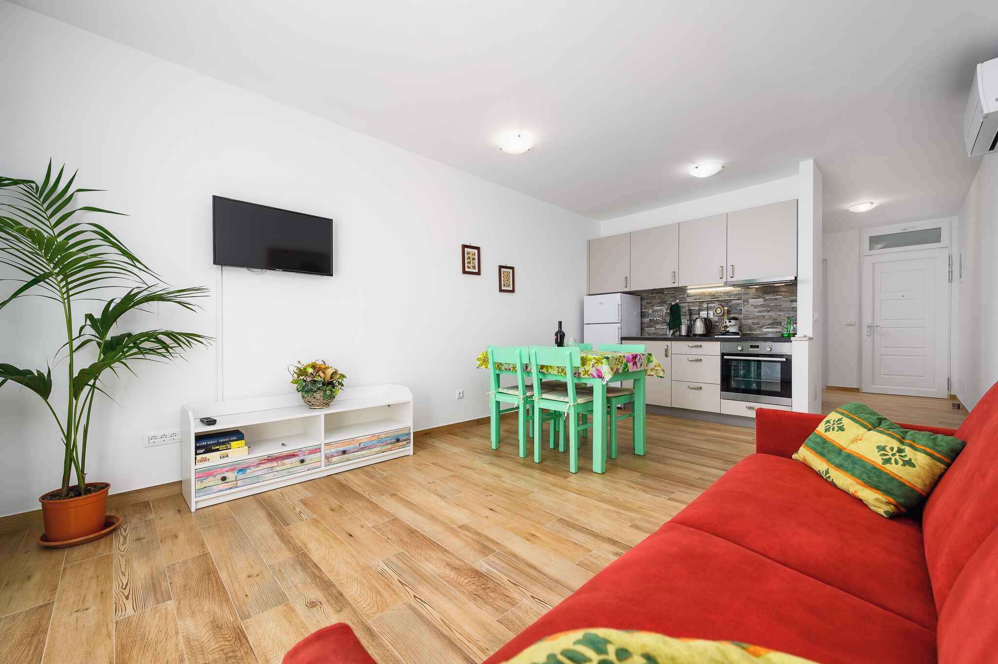 A modern living room with a red sofa, a dining table with green chairs, a TV mounted on the wall, and a kitchen area with white cabinets.