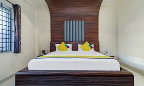 Una habitación de hotel con una cama grande, un cabecero de madera, una ventana con cortina y una pequeña mesa con una botella de agua y un teléfono.