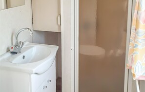 Kylpyhuoneessa on valkoinen pesuallas, peili ja ovi, joka johtaa suihkutilaan.