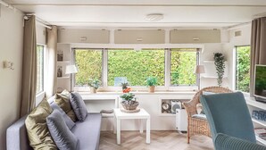 Een gezellige woonkamer met een bank, een kleine tafel en een stoel. Er zijn ramen met planten op de vensterbank en uitzicht op groen buiten.
