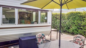 Een terras met een tafel en stoelen, een gele parasol, en een witte camper met 'SANTANA' erop.