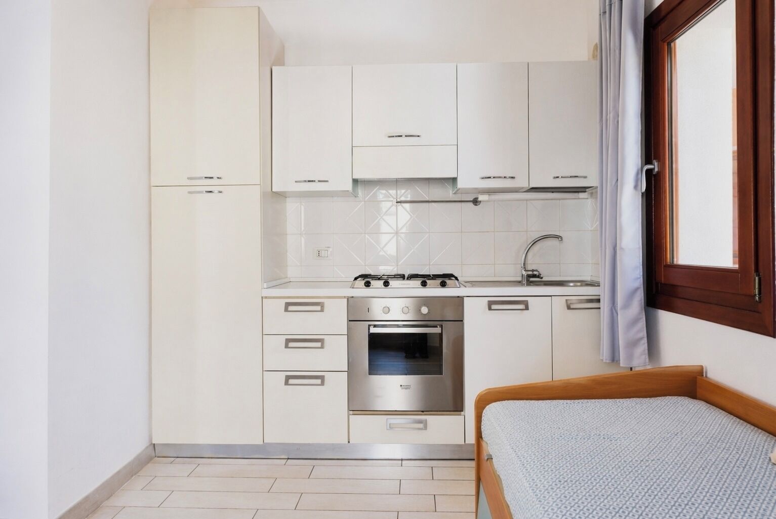 Una cocina compacta con armarios blancos, estufa, horno y una mesita de noche junto a la cama.