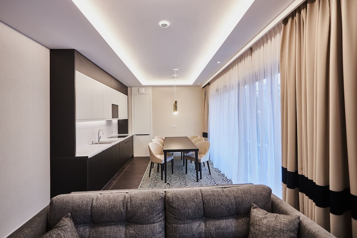 Una sala moderna con sofá, un comedor con mesa y sillas, una cocina con pileta y alacenas, y amplias ventanas con cortinas.