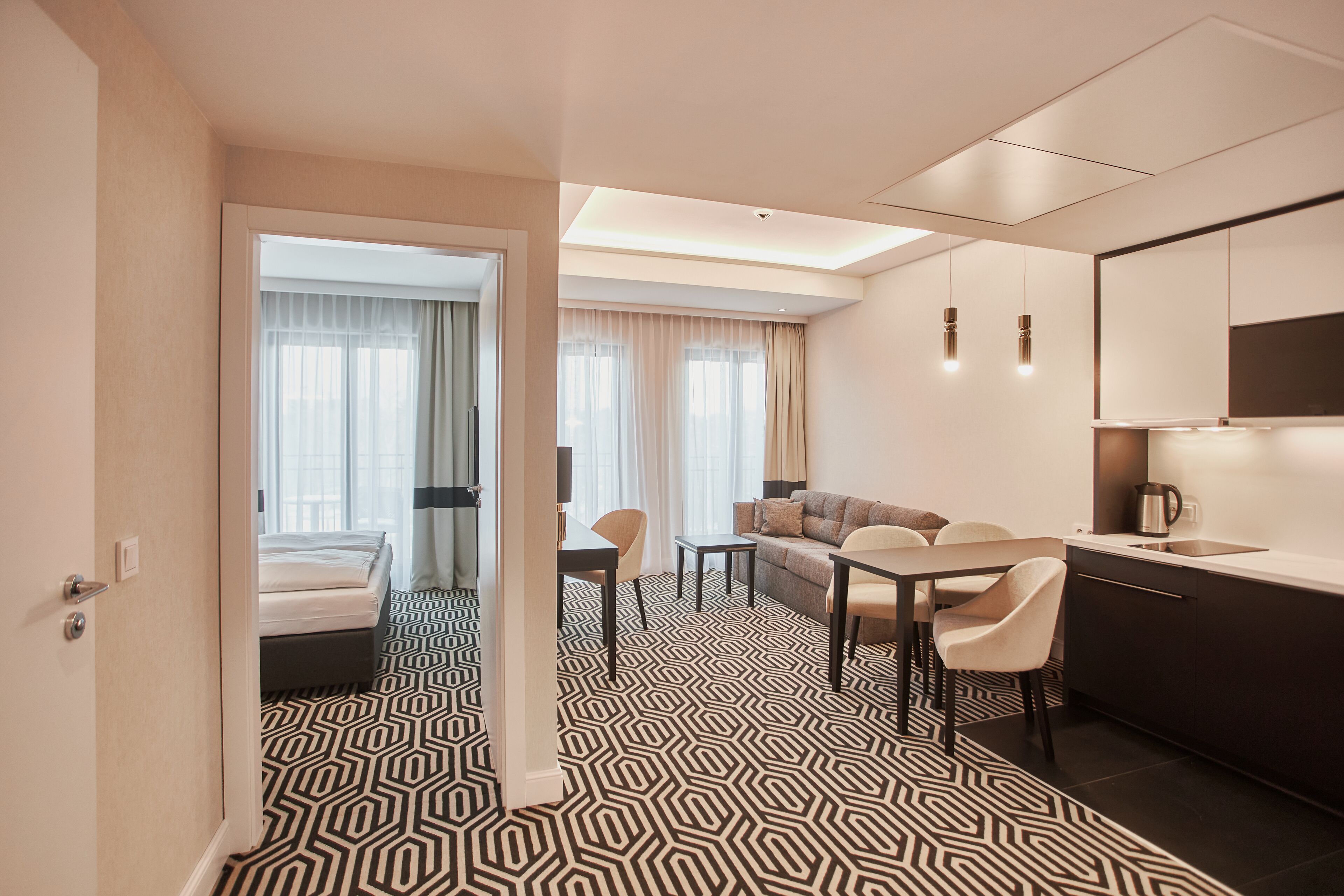 A modern hotel room with a geometric patterned floor, a sofa, a dining table, and a kitchenette.