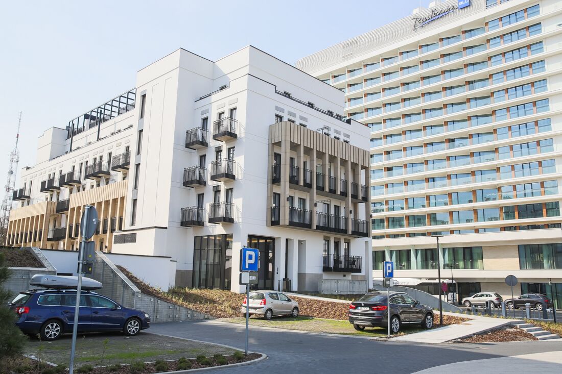 Een modern gebouw met een witte gevel, meerdere balkons en een parkeerplaats met aangegeven parkeerplaatsen.