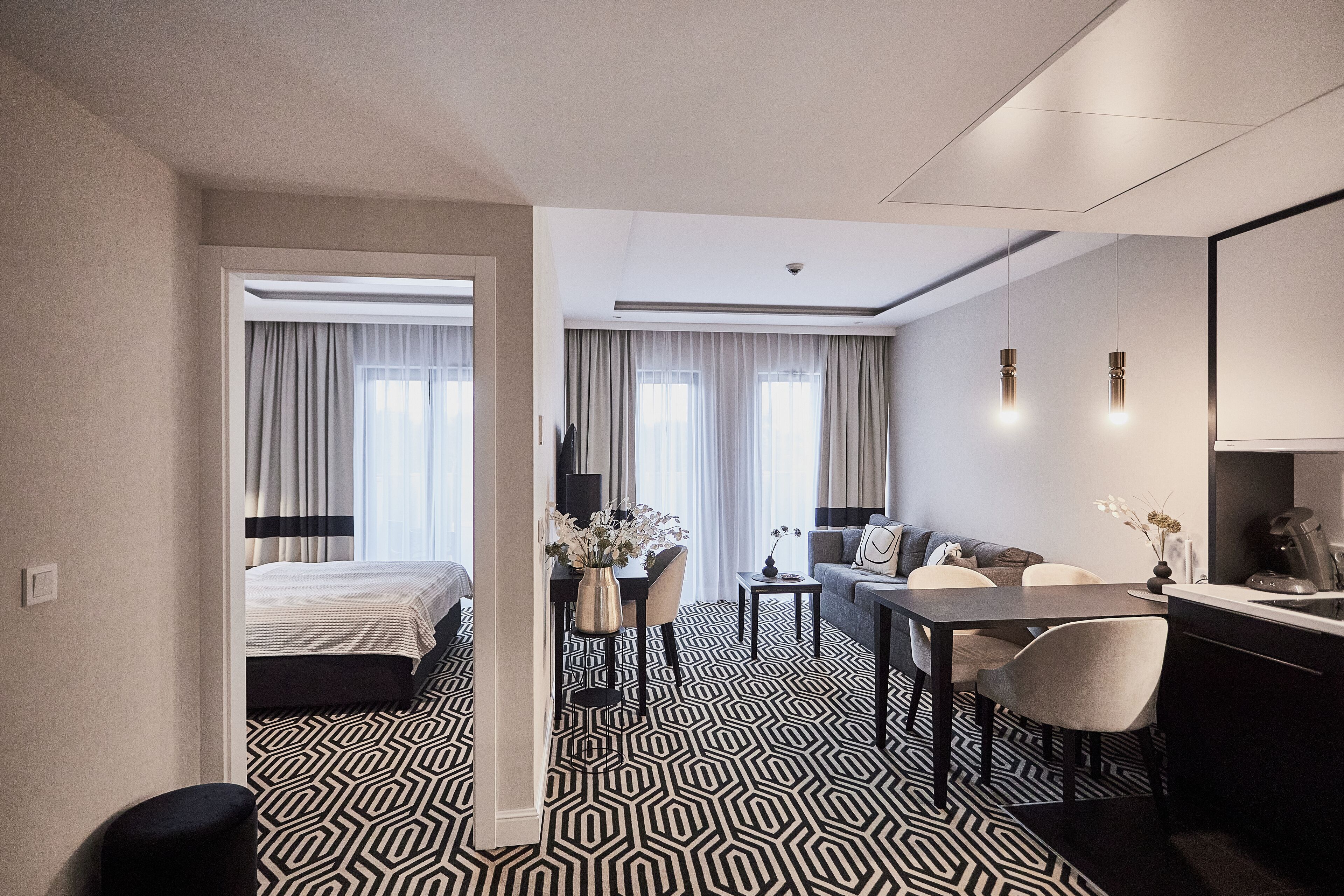 Modernilla designilla sisustettu hotellihuone, jossa on suuri sänky, työpöytä, sohva ja ruokailutila. Huoneessa on geometrisesti kuvioitu lattia ja suuret ikkunat, joissa on verhot.