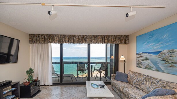 Een woonkamer met een grote televisie, een bank, een salontafel en een balkon met uitzicht op de oceaan.