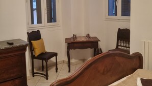 A room with a bed, a wooden dresser, a chair, a small table with a chessboard, and two windows with blinds.