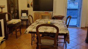 A dining area with a tiled floor, a table with a patterned tablecloth, wooden chairs, a television on a stand, and a cabinet with glass doors.