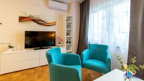 Un salón moderno con un televisor de pantalla plana, sillas color turquesa, una mesa consola blanca y un amplio ventanal con cortinas traslúcidas.