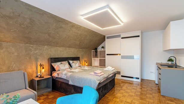 Un dormitorio moderno con cama, mesitas de noche, una silla, armario y kitchenette.