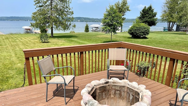 Une terrasse en bois avec un foyer et deux chaises, donnant sur un lac où l’on peut voir des bateaux et une zone herbeuse.