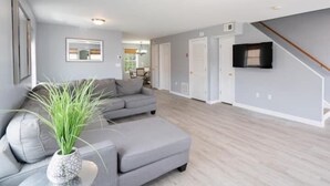 A modern living room with a grey sofa, a small table with a plant, and a staircase leading to another level. - Gorgeous Waterfront 3 Bedroom, 1.5 Bathroom Unit. (New Haven)
