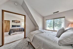 Un dormitorio con un ventanal grande, una cama con cubrecama gris y un espejo que refleja la habitación.