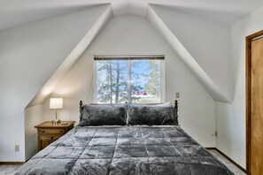 A bedroom with a sloped ceiling, a large window, a bed with a dark patterned bedspread, a bedside table with a lamp, and a wooden door.