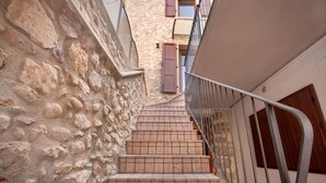 Un escalier étroit en pierre mène à une porte en bois, flanquée d’une rambarde en métal et d’un mur en pierre.