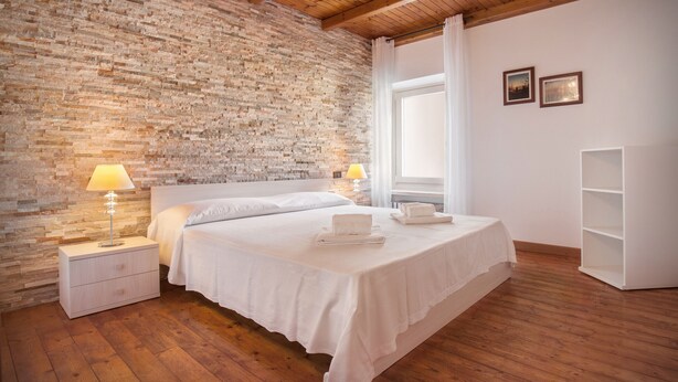 Une chambre avec un mur en briques, un plancher en bois, un lit blanc, des tables de chevet avec des lampes et une fenêtre avec des rideaux.