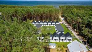 A row of white cabins with dark roofs, surrounded by dense pine trees, and a view of the sea in the background.