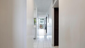 A narrow hallway with white walls and a tiled floor, leading to a room with a window and a door. - Summer Deal Downtown Kona Vacation Condo Walk to Shops Dining and Beach (Kailua Kona)