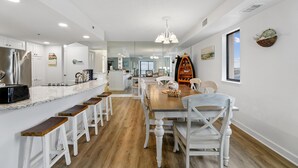 A modern kitchen with white cabinetry, a wooden dining table, and bar stools. - DAILY ACTIVITIES & LINENS INCLUDED*! CAPRI - 3BR Direct Oceanfront Updated Condo! (Ocean City)