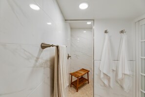Une salle de bain aux murs recouverts de marbre blanc, avec un tabouret en bois et deux serviettes blanches suspendues à des crochets.