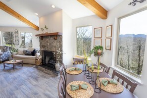 Una sala de estar acogedora con chimenea de piedra, una zona de comedor con individuales tejidos y un gran ventanal con vistas a árboles.