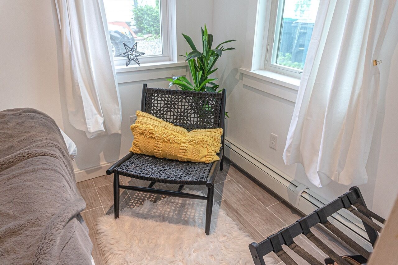 A corner room with a black wicker chair, a yellow cushion, a white furry rug, and a potted plant by a window.